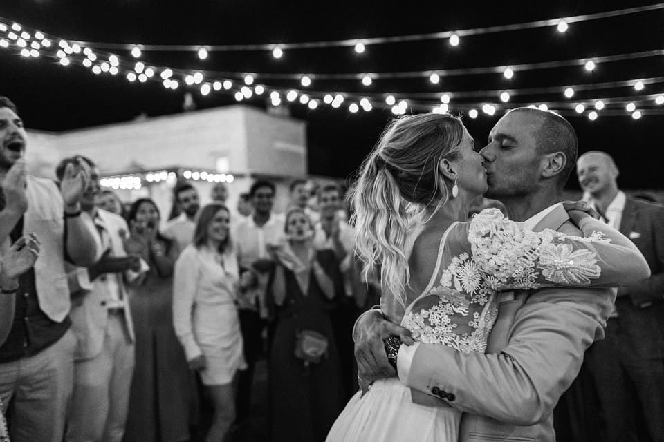 Une photo en noir et blanc. Elle est prise de nuit et montre deux mariés s'embrassant tendrement entourés de leurs amis et de leur famille.