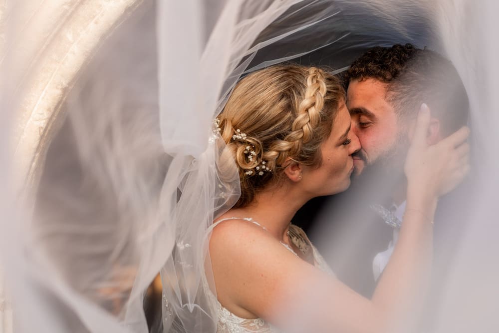 Couple de mariés en séance day ater qui s'embrassent, le voile qui s'envole forme un halo autour de leurs deux visages